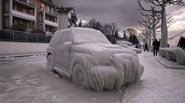 A car is completely encased in thick, textured ice and snow, with icicles hanging from its body, parked on a snowy street…