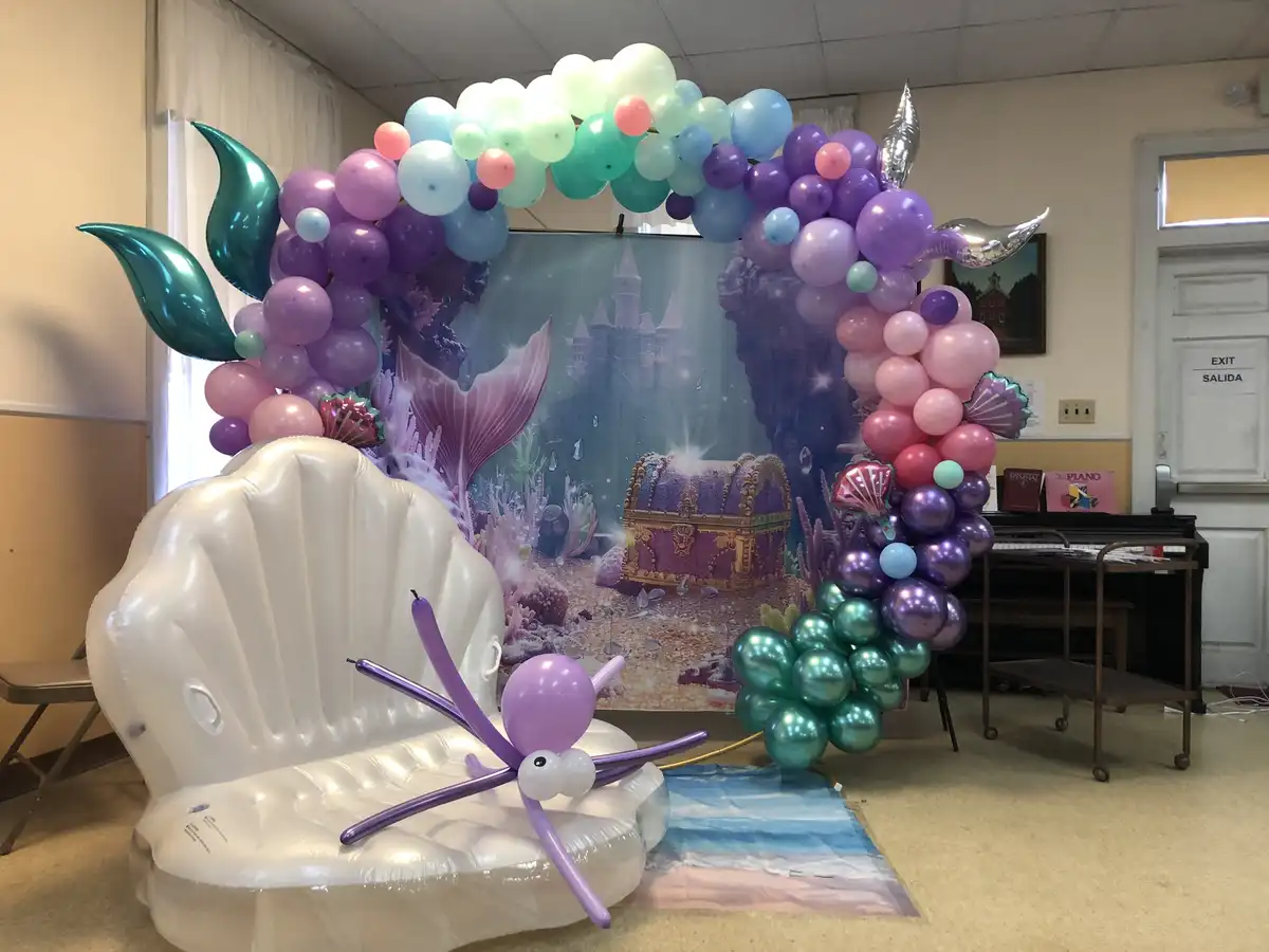 The image shows a mermaid-themed party setup in a room with a large inflatable seashell chair in the foreground. A balloon…