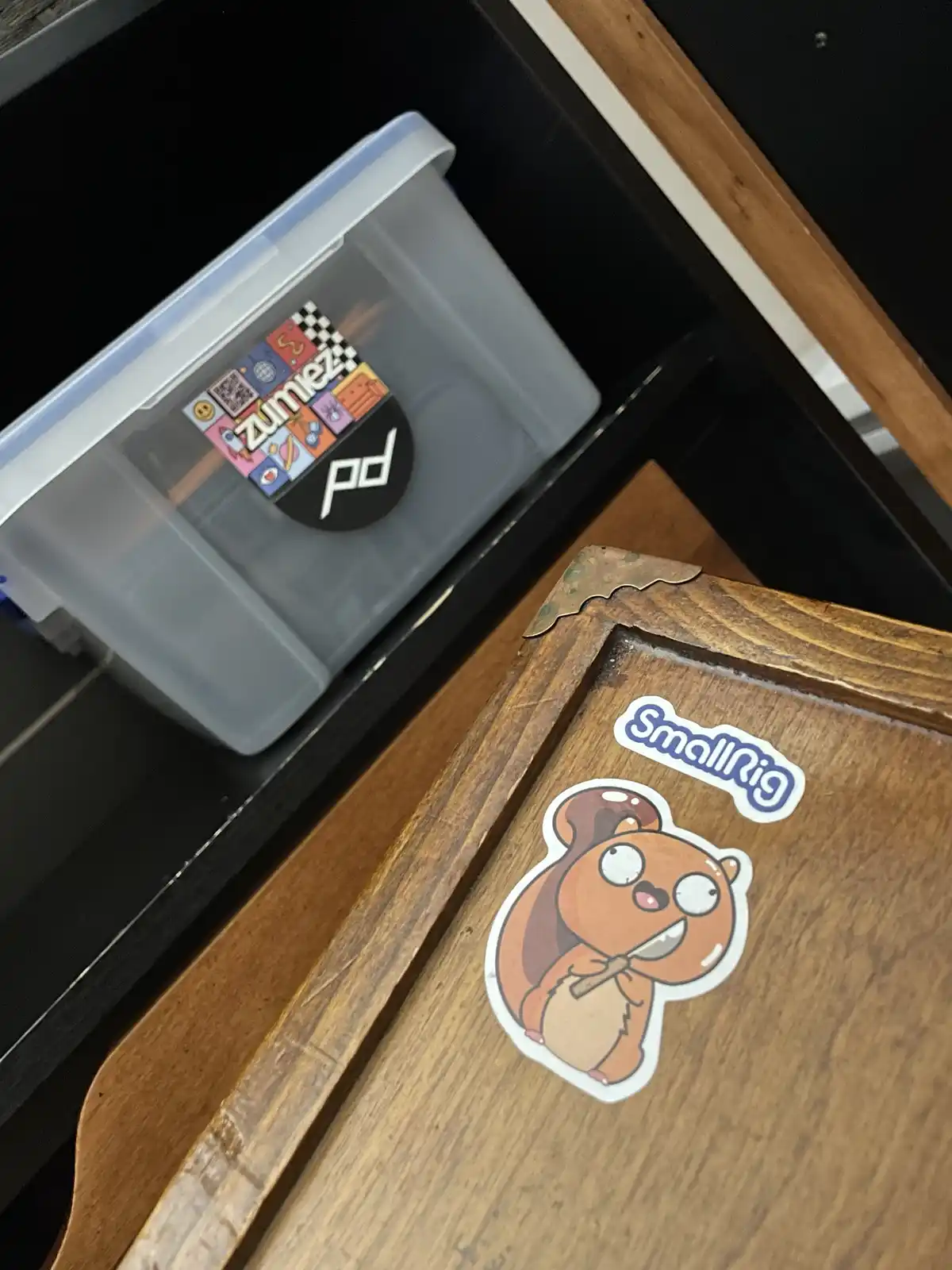 A close-up photo shows a wooden surface with a cartoon squirrel sticker labeled 'SmallRig' and a clear plastic storage bin…