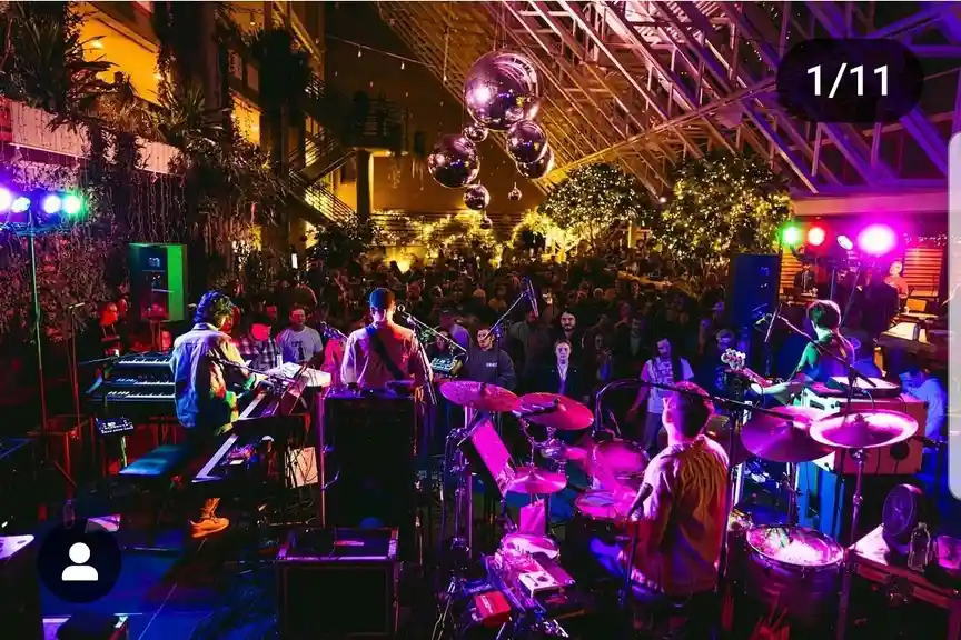 A vibrant, crowded indoor concert scene with a band performing on stage under colorful stage lights and hanging disco balls…