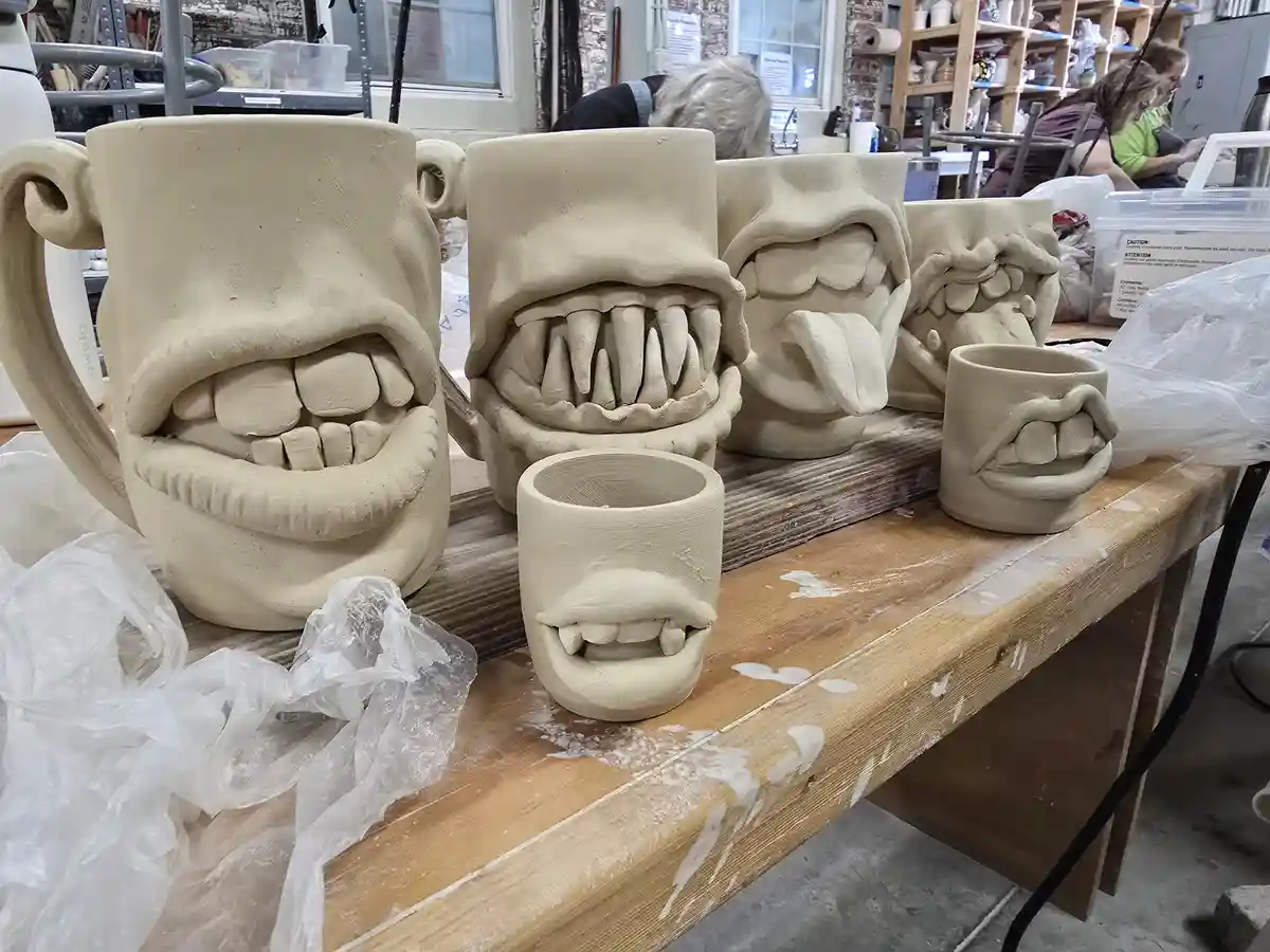 A row of unfinished clay mugs, each shaped like a grotesque, exaggerated human face with wide-open mouths and teeth, sits on…
