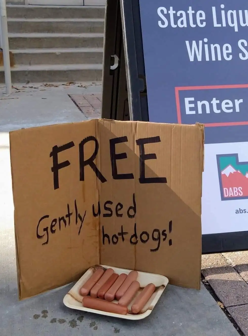 A handmade cardboard sign on a sidewalk reads 'FREE Gently used hotdogs!' and sits beside a styrofoam tray holding several…