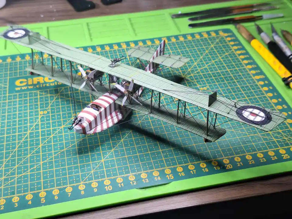 A detailed model of a biplane with green wings and a red-and-white striped fuselage sits on a green cutting mat with yellow…