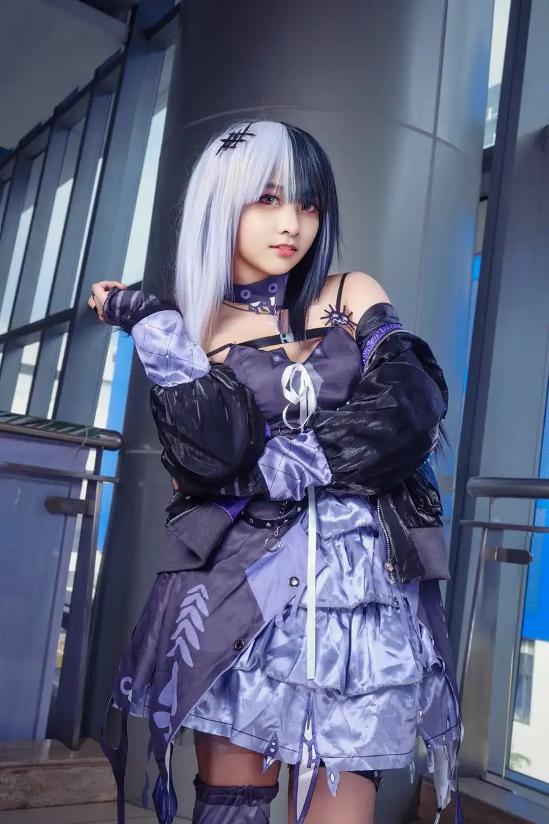 A person in a detailed purple and black cosplay outfit with silver and black hair, standing indoors near glass walls. The…