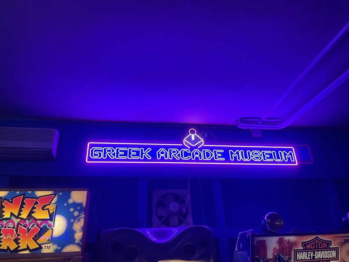 A neon sign glowing purple and white reads 'GREEK ARCADE MUSEUM' above arcade machines in a dimly lit room bathed in blue…