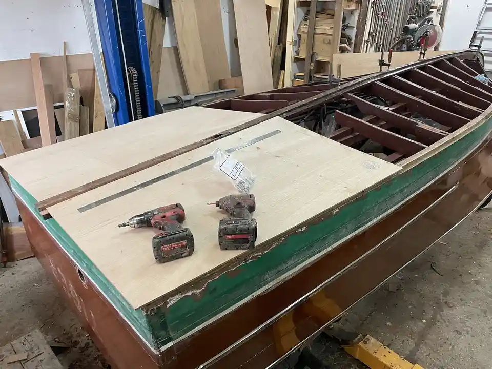 A partially built wooden boat sits in a workshop, its hull painted green and brown with visible wear. On top of the boat's…