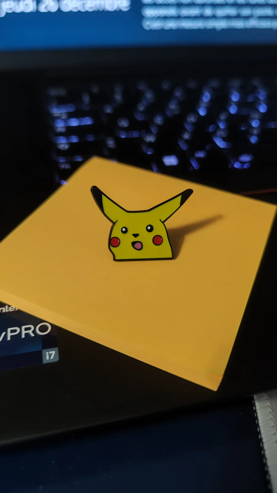 A yellow Pikachu enamel pin with a surprised expression sits on a yellow sticky note placed on a laptop keyboard. The laptop…