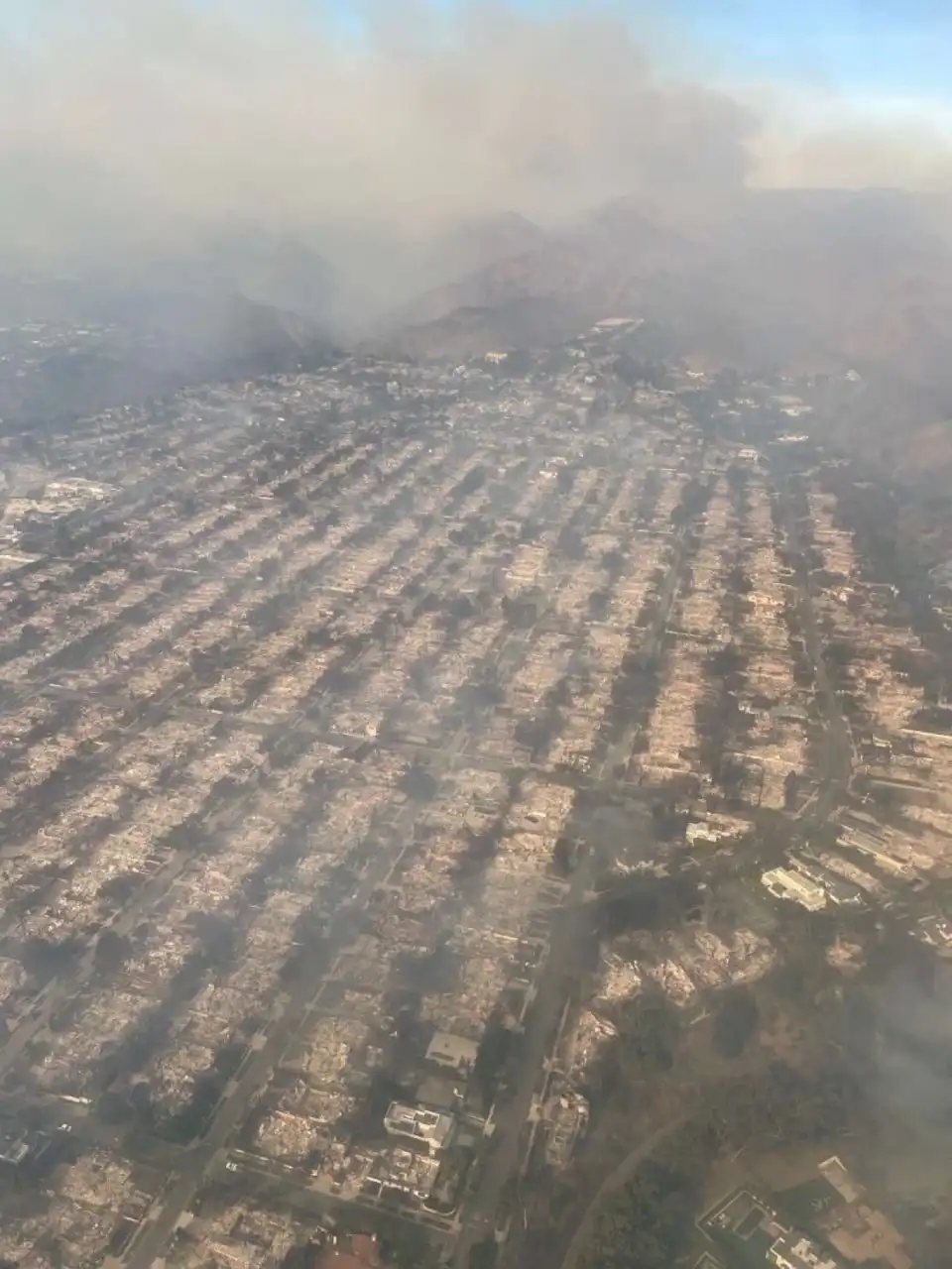 An aerial view shows a vast suburban area completely destroyed by fire, with charred remains of houses and streets, heavy…