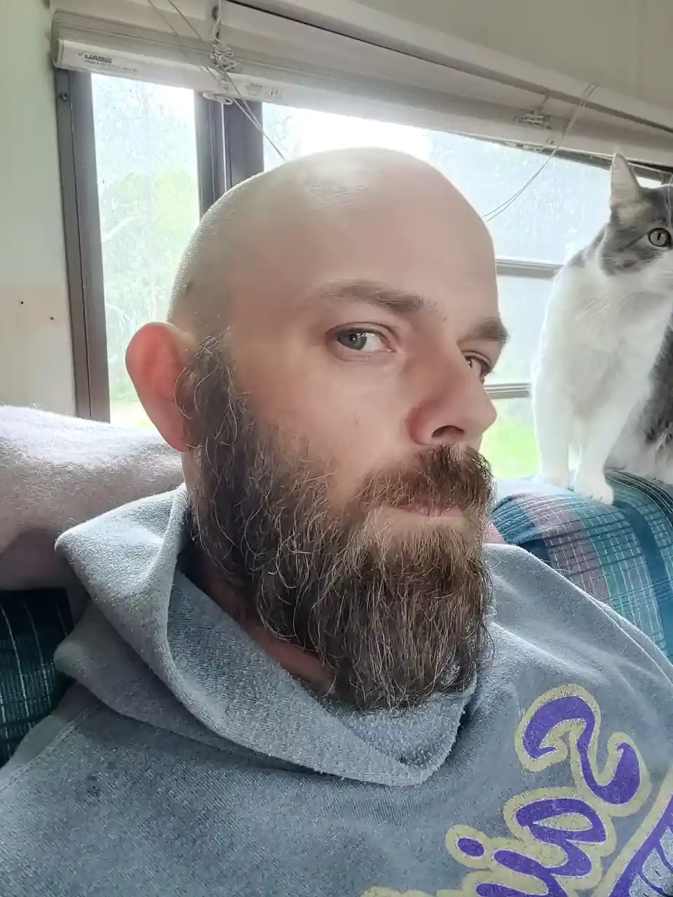 A bald man with a full beard wearing a grey hoodie sits on a couch, looking slightly to his right with a neutral expression…