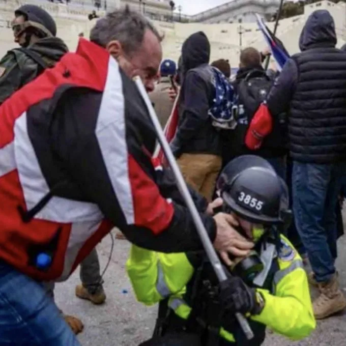 A man in a red and black jacket aggressively grabs the face of a police officer in a yellow vest and black helmet marked…