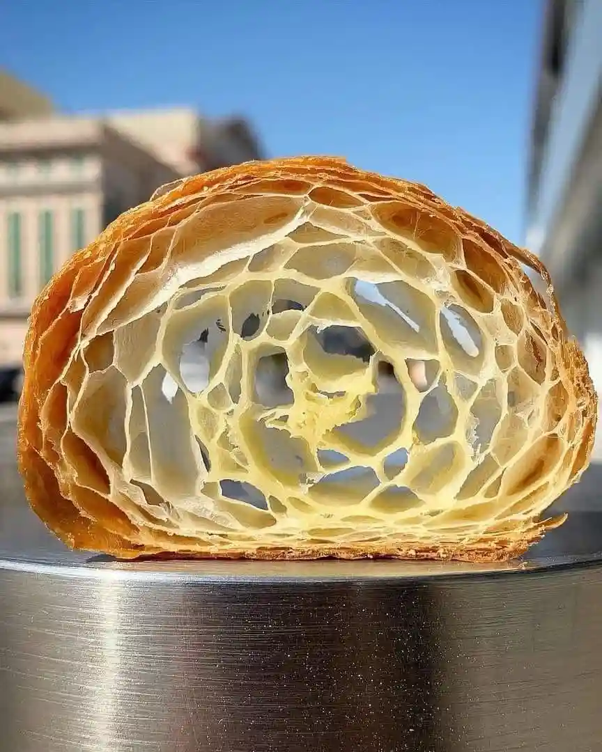 A close-up photo shows a sliced croissant revealing its intricate, honeycomb-like flaky interior with golden-brown crust…