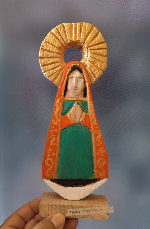 A hand holds a small, hand-carved wooden statue of a religious figure resembling the Virgin Mary, featuring a gold halo, an…
