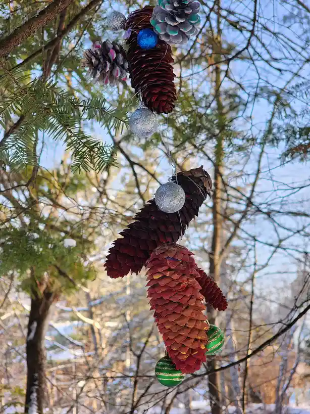 A pinecone is decorated with colorful ornaments and strung with lights, hanging from a tree branch in a snowy forest…