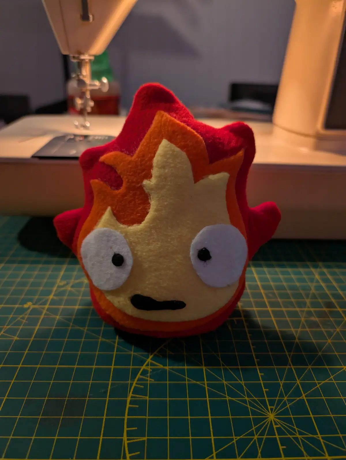 A handmade felt plush toy shaped like a cartoon fireball with red, orange, and yellow layers, large white eyes with black…