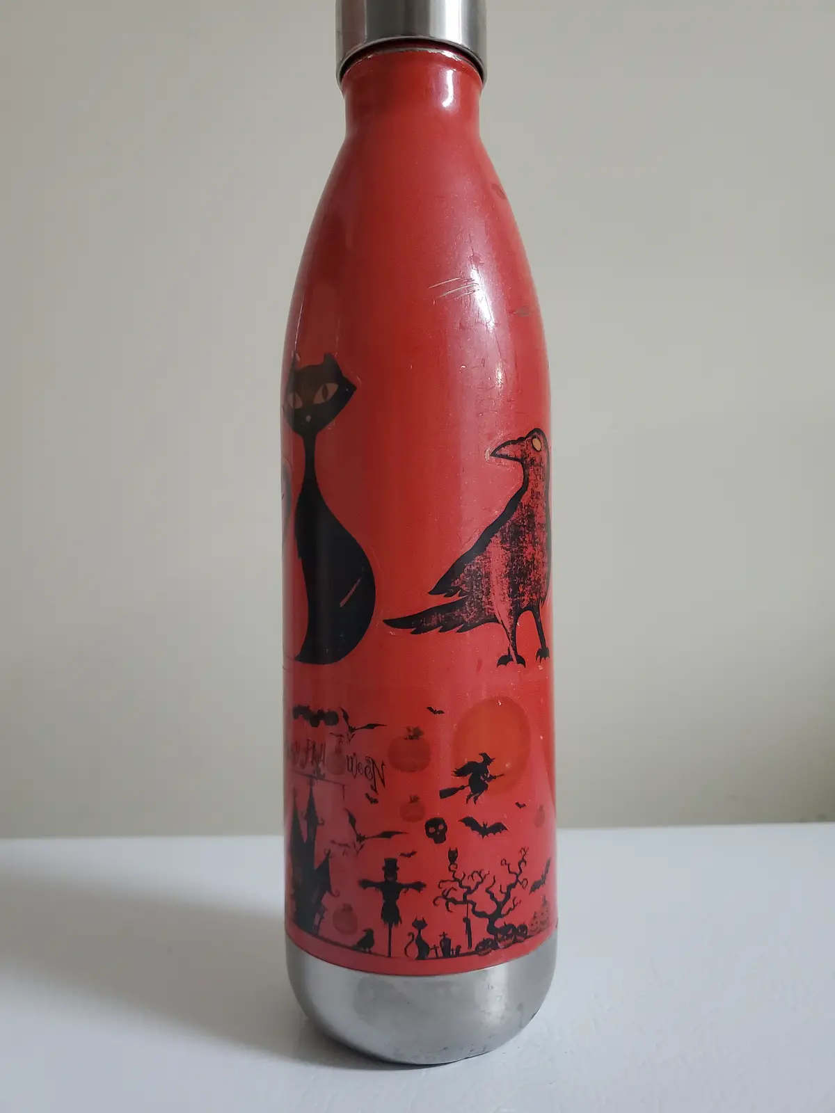 A red reusable water bottle with a silver cap and base, featuring a Halloween-themed design with black silhouettes of a cat…
