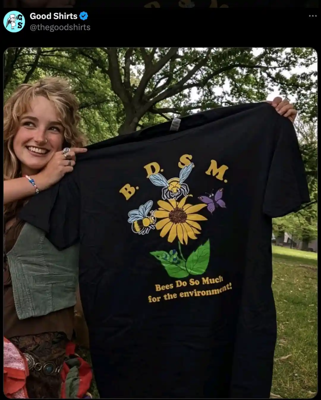 A smiling woman holds up a black t-shirt outdoors that features a graphic of bees, a butterfly, and a sunflower. The shirt…