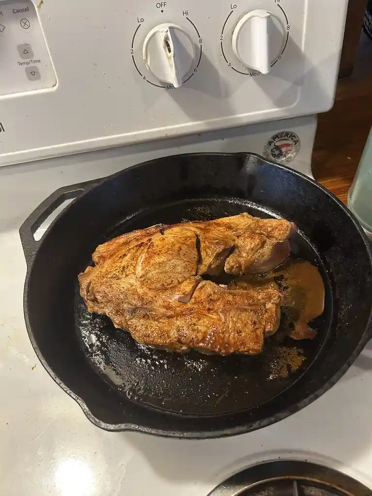 A close-up photo shows a large, seared piece of meat cooking in a black cast-iron skillet on a white electric stove. The…