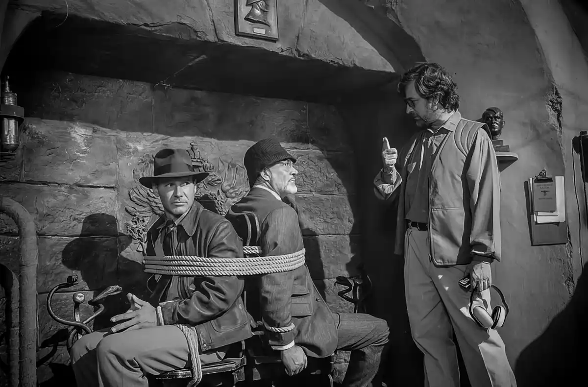 A black-and-white photo shows two wax figures of Indiana Jones and another man, both bound with rope in a stone-walled room…