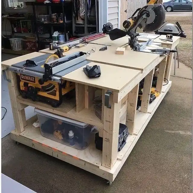 A custom-built wooden workbench in a garage, featuring a yellow DeWalt table saw and a yellow DeWalt miter saw mounted on…