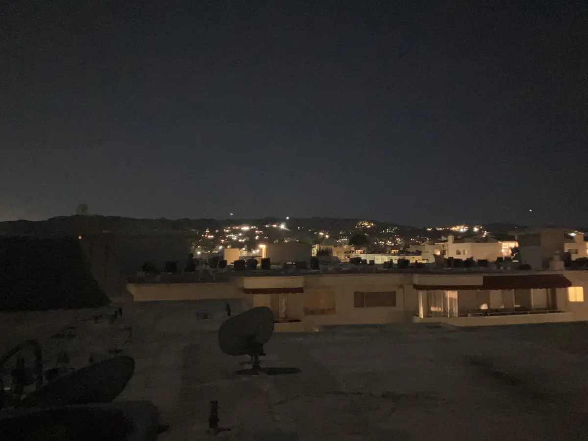 A nighttime rooftop view showing a satellite dish, distant city lights on hills, and buildings under a dark sky. The…