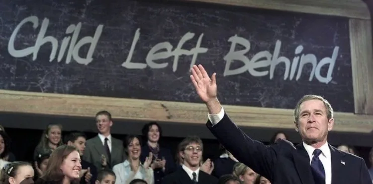 A man in a suit waves to a crowd while standing in front of a chalkboard that reads 'Child Left Behind'. The crowd includes…