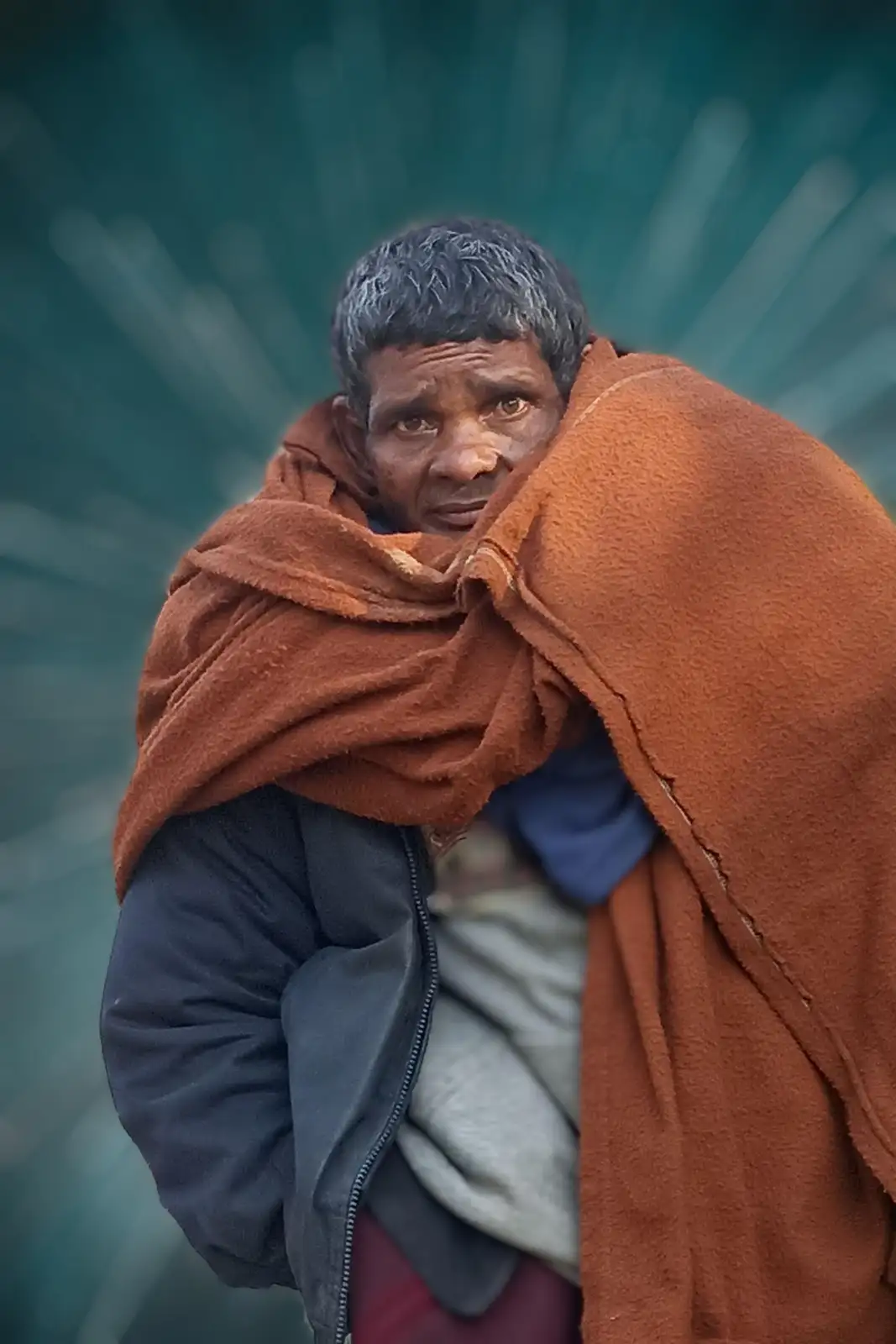 A man with dark, short hair and a weathered face is wrapped tightly in a worn, brown blanket or shawl, with a dark jacket…