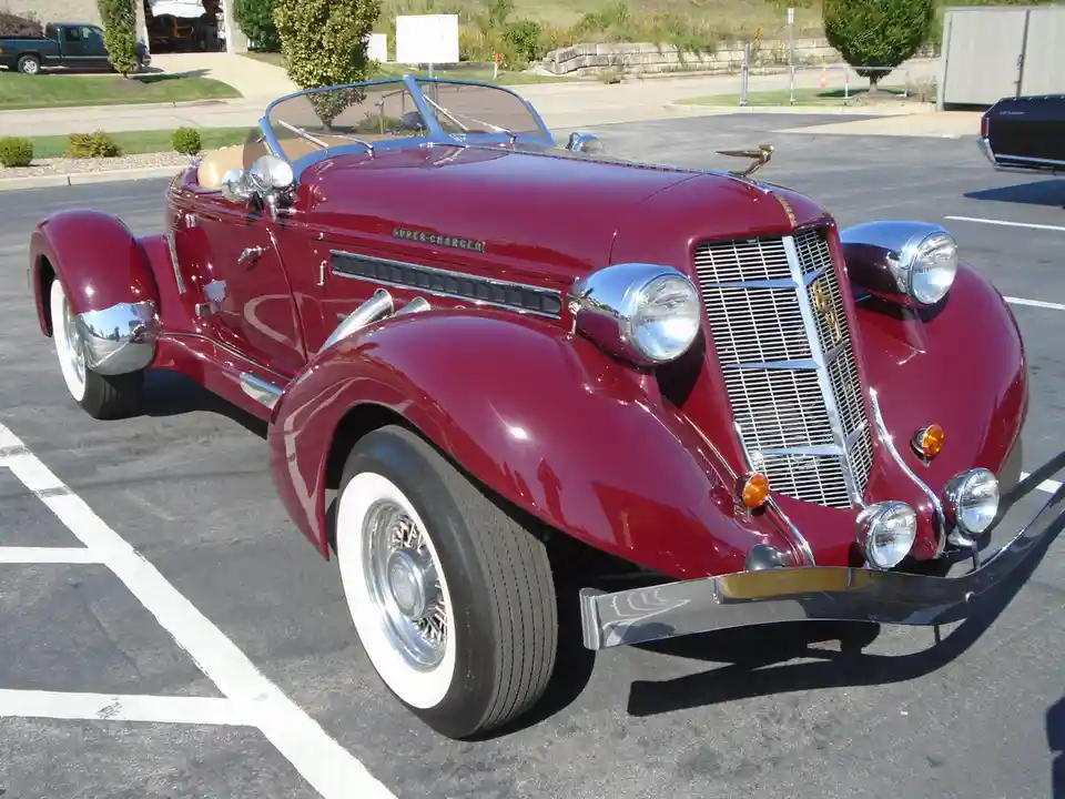 A shiny, maroon classic convertible car with chrome accents and white-wall tires is parked in a lot. The car's hood has a…