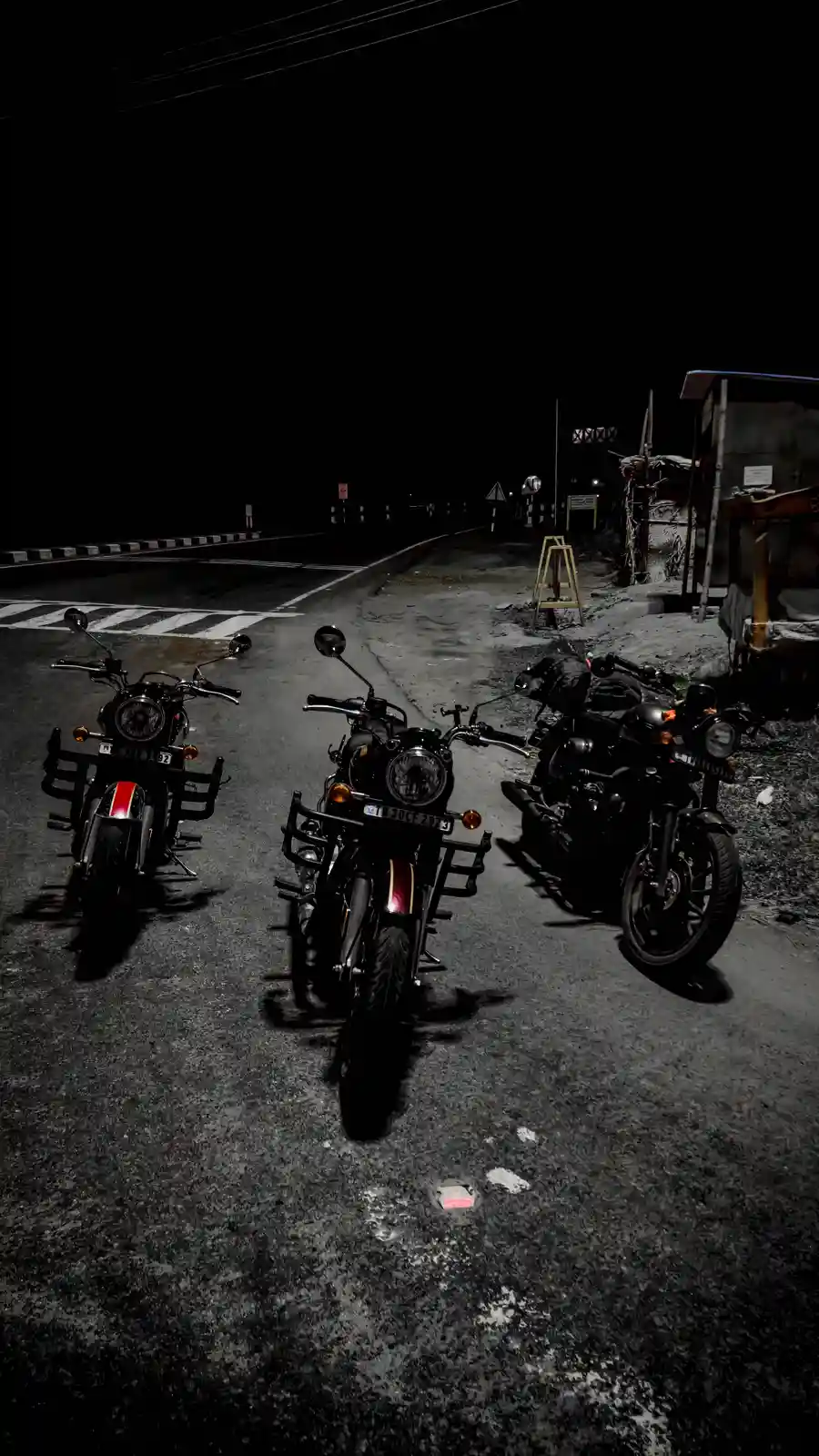 Three black motorcycles are parked on a dark, empty road at night, with a crosswalk visible in the background. The image is…