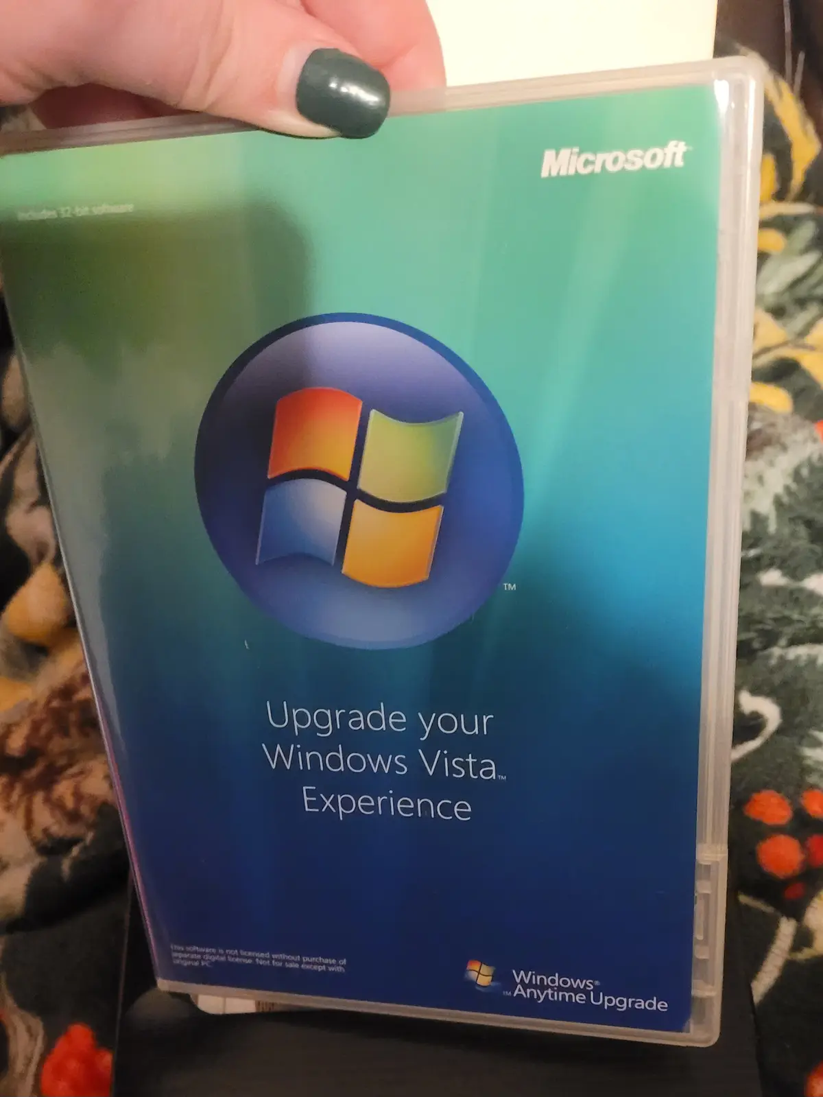A hand with dark green nail polish holds a Windows Vista Anytime Upgrade software box. The box features the classic Windows…