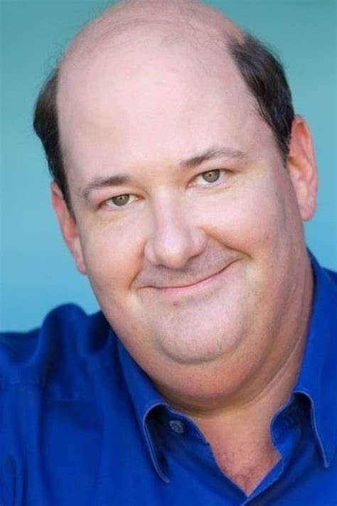 A man with a balding head and a subtle, smug smile wears a blue collared shirt against a solid blue background. The overlaid…