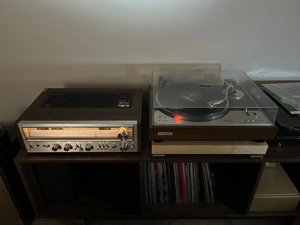 A vintage Pioneer stereo receiver and turntable sit side-by-side on a wooden shelf, with a stack of vinyl records visible…