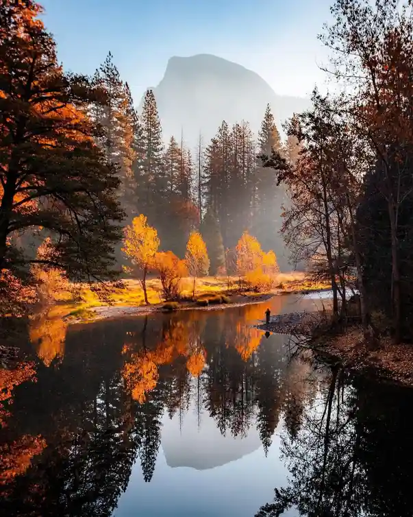 A serene autumn landscape featuring a calm river reflecting a mountain and trees with golden foliage, with a small human…