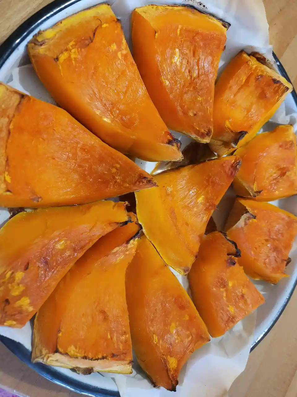 A white enamel bowl with a black rim holds roasted butternut squash wedges on parchment paper. The squash is bright orange…