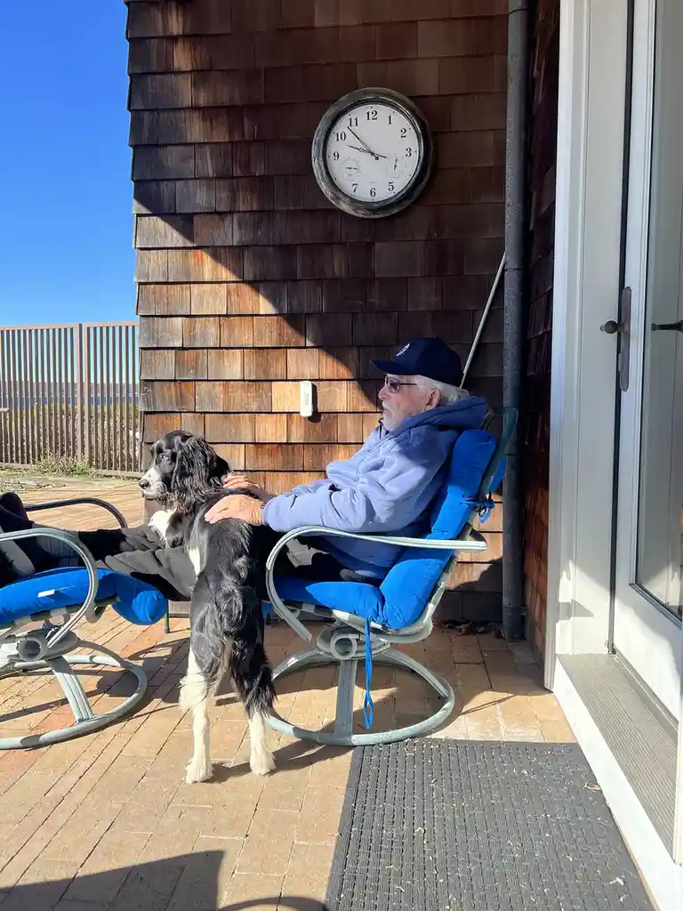 An older man with white hair and glasses sits in a blue outdoor chair on a wooden deck, petting a black and white dog…