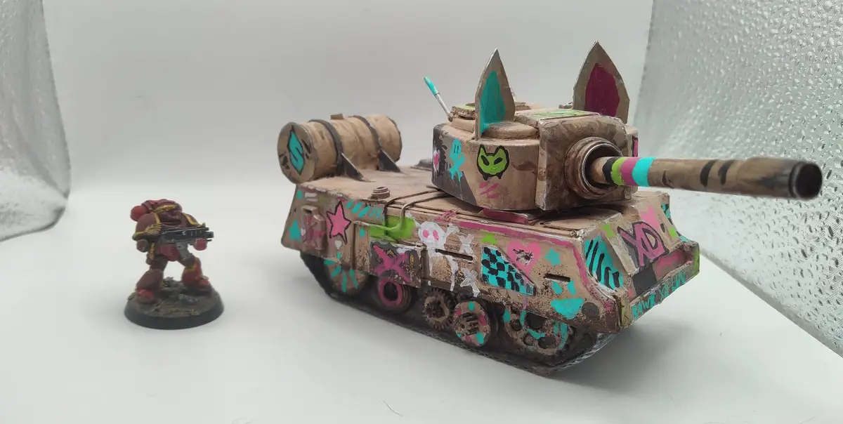 A small, red-painted miniature warrior figure stands beside a heavily customized, brown tank model decorated with colorful…