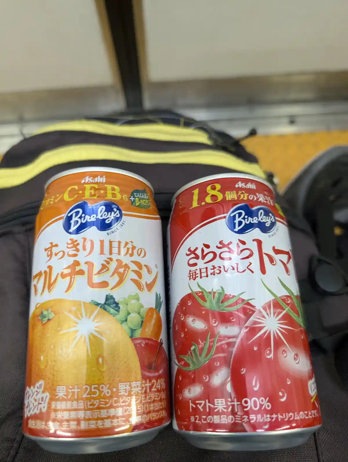 Two Asahi Bireley's canned drinks sit side by side on a black and yellow striped bag. The left can is orange and labeled…