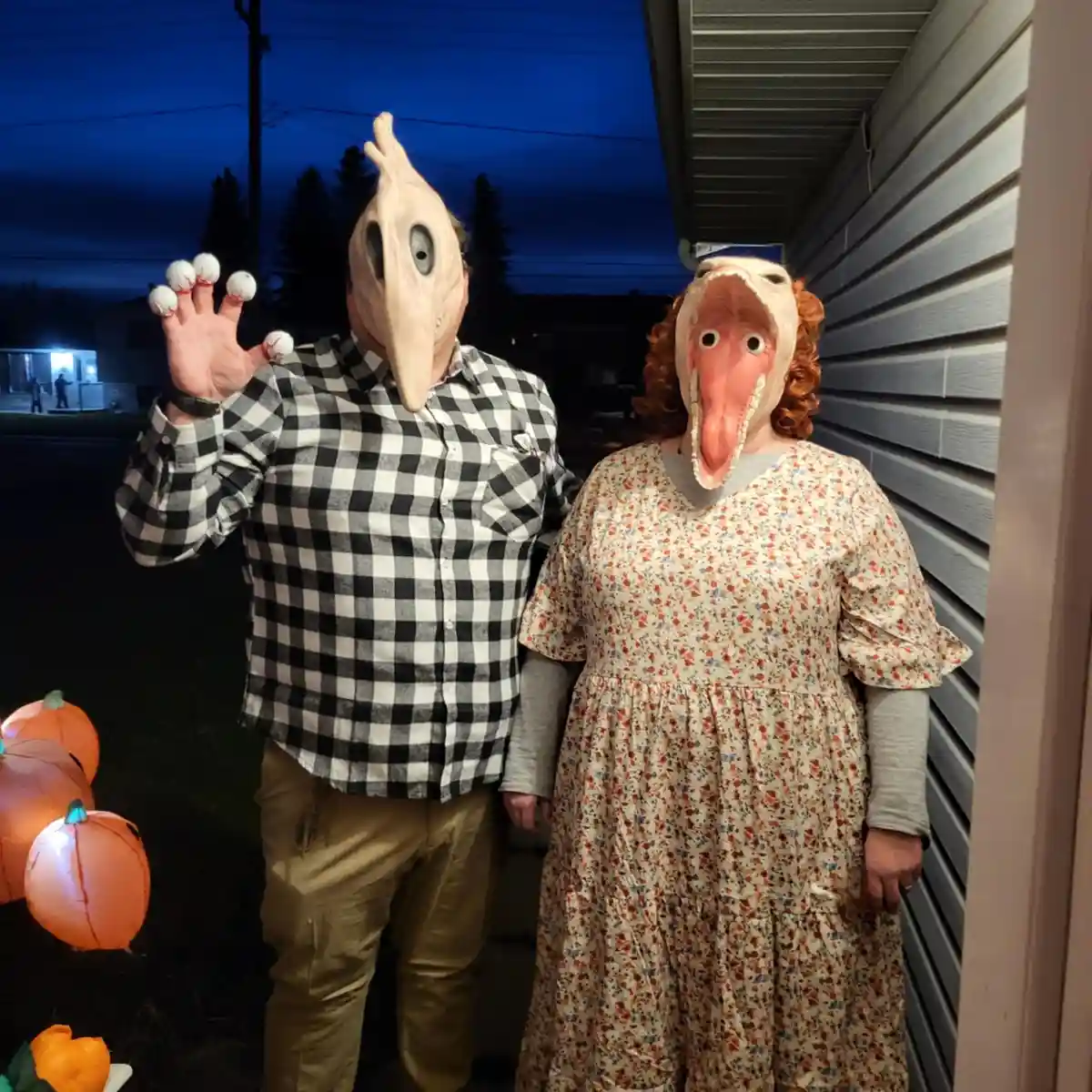 Two people stand outside a house at night, both wearing large, grotesque bird masks. The person on the left, in a plaid…