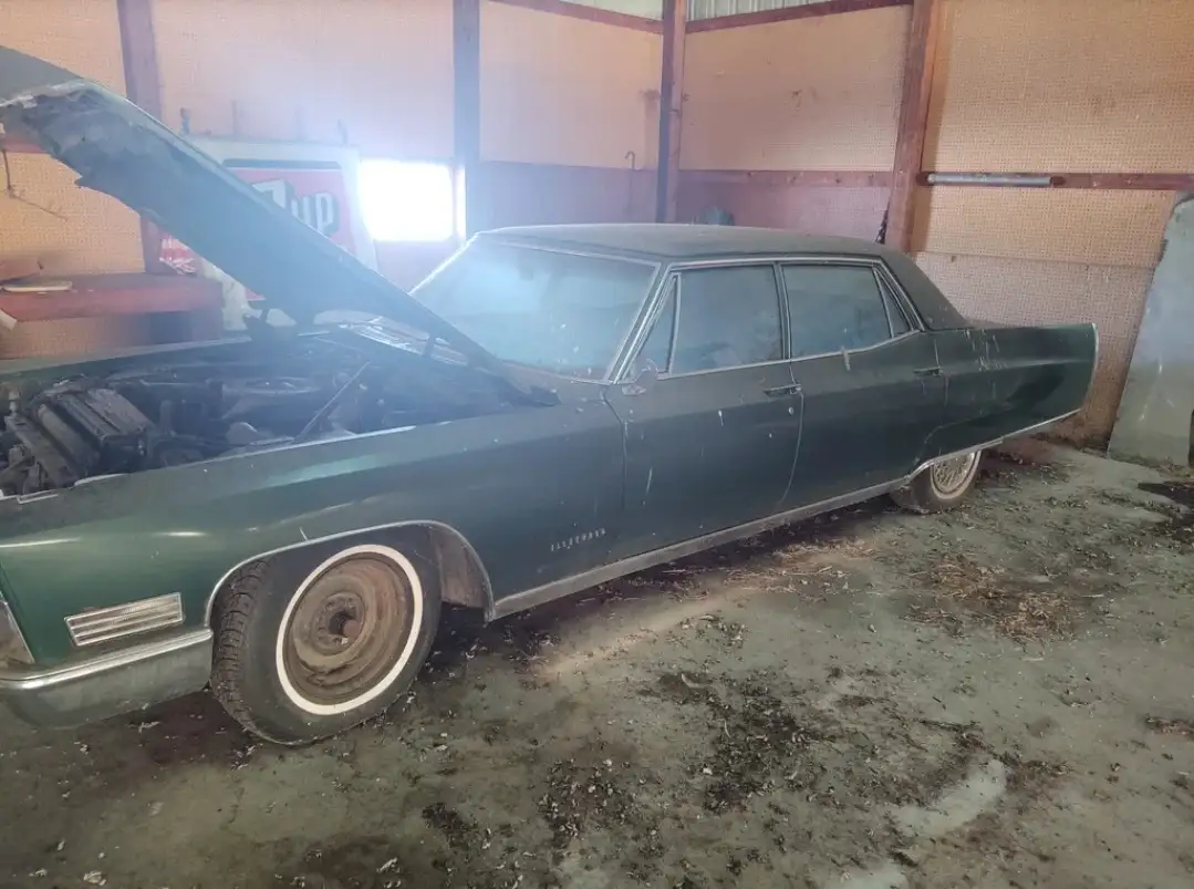 A dusty, green classic car with its hood open sits inside a cluttered, dimly lit barn. The vehicle appears neglected, with…