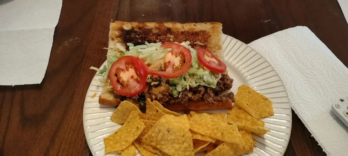 A close-up photo shows a sandwich on a white paper plate, made with toasted bread, ground meat, shredded lettuce, and tomato…