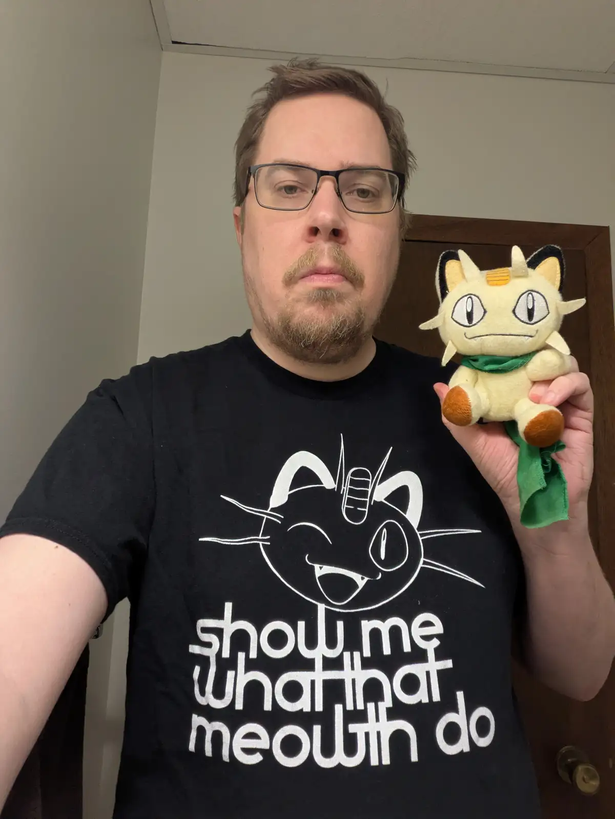 A man with glasses and a goatee wears a black t-shirt with a white graphic of a cat and the text 'show me what that meowth…