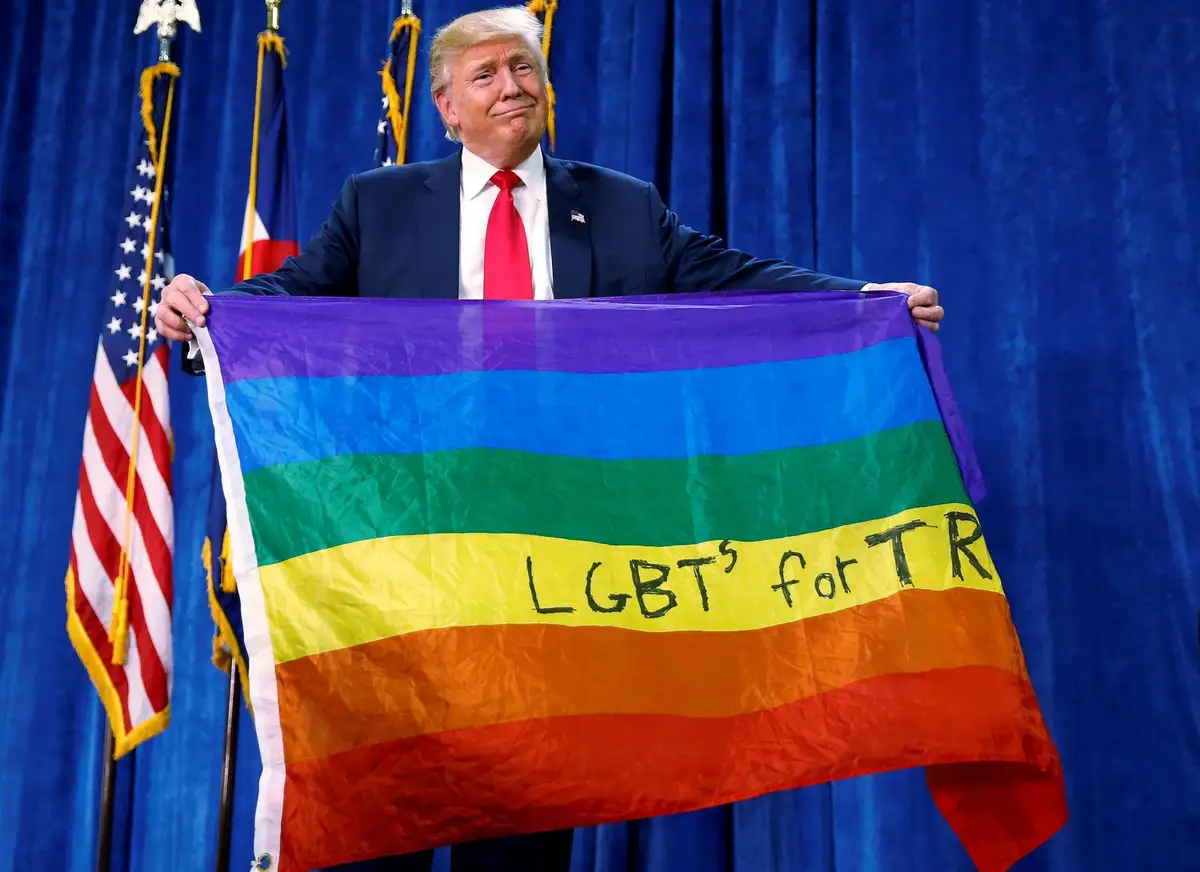 A man in a suit and red tie stands in front of blue curtains and American flags, holding a rainbow flag with the text…