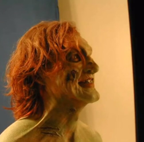 A person with pale, greenish-gray makeup resembling a zombie or monster, messy reddish-brown hair, and a wide, unsettling…