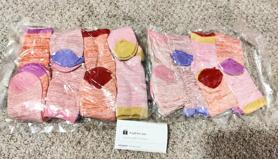 Two clear plastic packages of colorful, patterned socks lie on a beige carpet, each containing multiple pairs with…