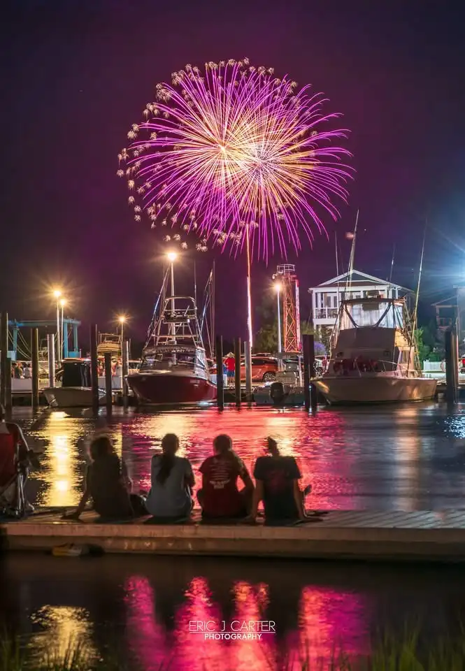 Four people sit on a dock at night watching a large, colorful fireworks display over a marina with boats and lights…