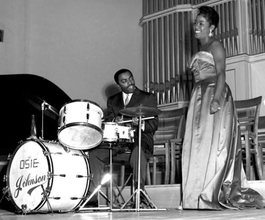 A black-and-white photo shows a man in a suit seated at a drum kit with 'OSIE JOHNSON' written on the bass drum, smiling…