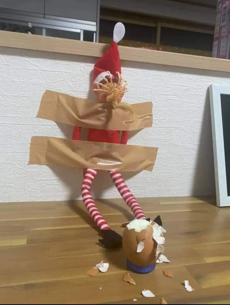 A red-clothed elf on the shelf is taped to a wall with brown duct tape, its head replaced by a cracked eggshell with a small…