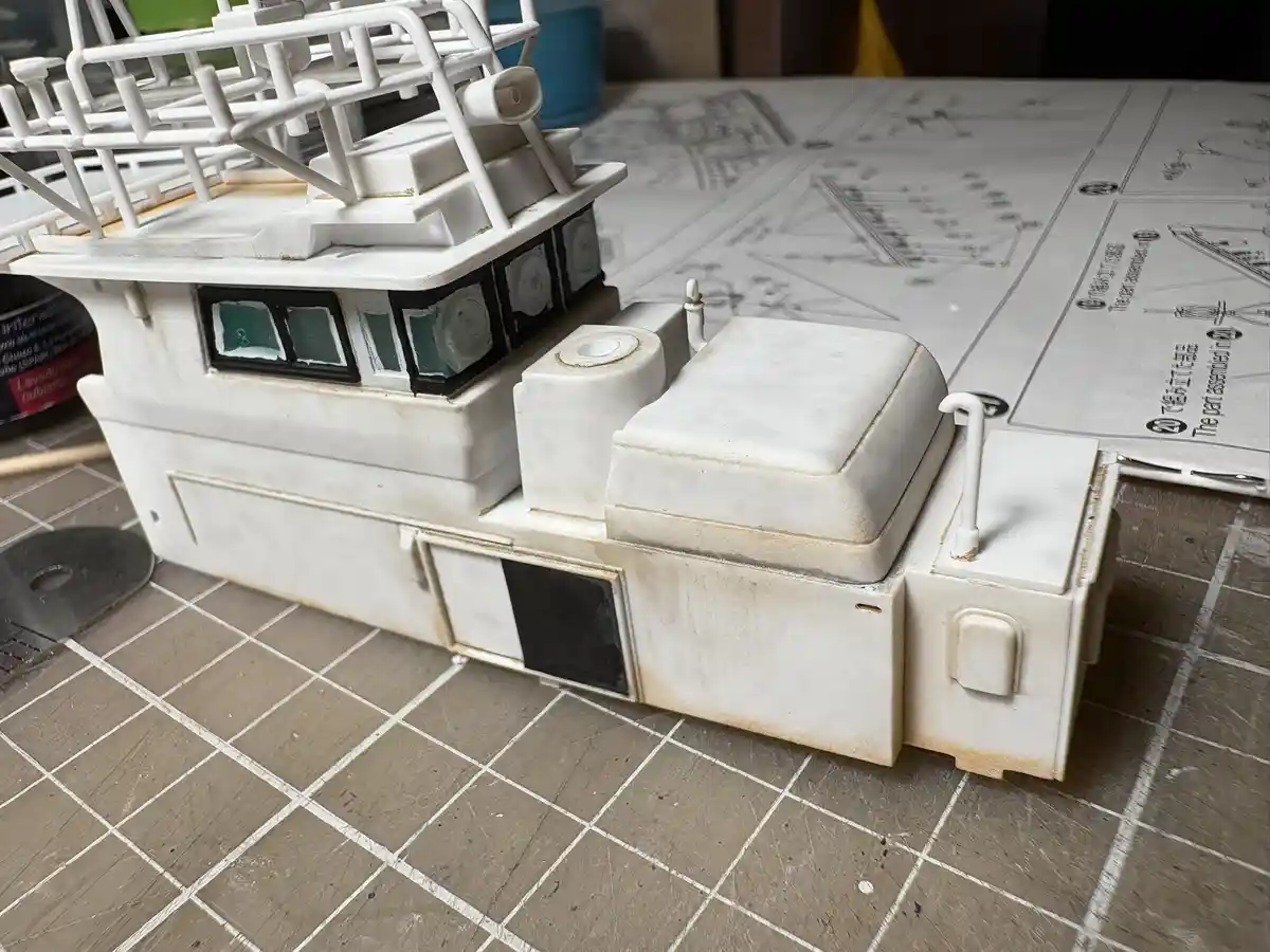 A partially assembled white plastic model ship sits on a gridded work surface next to its instruction manual, which shows…