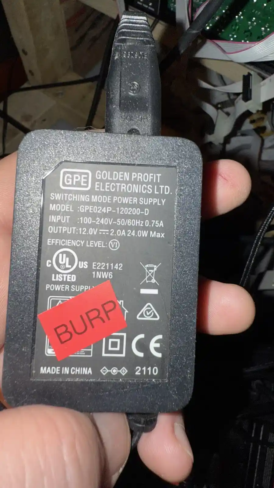A hand holds a black GPE brand switching mode power supply with visible technical specs and certification logos. A red…