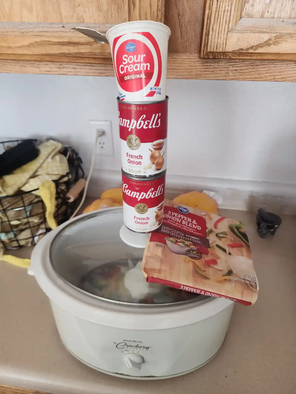 A white slow cooker sits on a kitchen counter with a stack of three food items balanced on its lid: a Kroger sour cream cup…