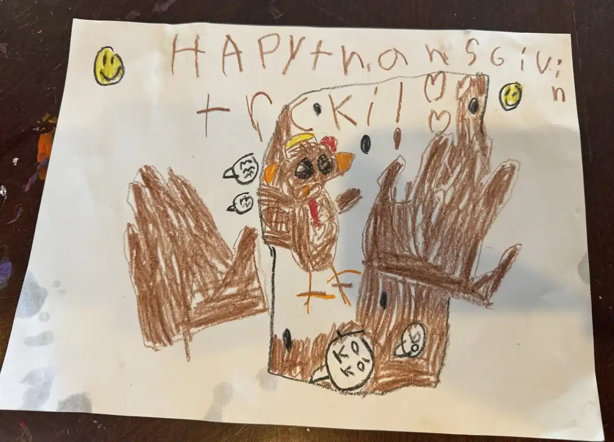 A child's crayon drawing on white paper shows a turkey with a red wattle and yellow beak, flanked by two brown handprints…